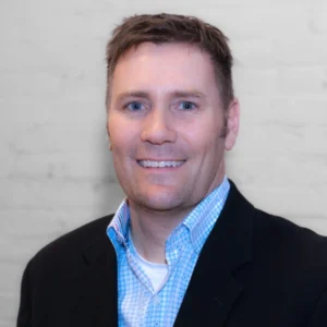A headshot of a man in a blue checkered shirt and black jacket