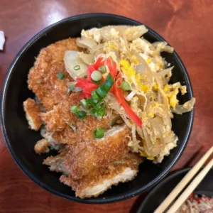 A black bowl with breaded chicken, green onions, and cabbage next to choptsticks
