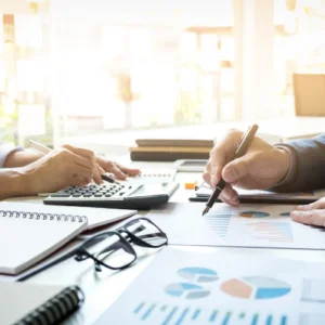 The hands of two business people are visible over notepads, calculators, glasses, and charts