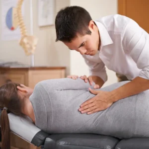 A male therapist presses into the back of a female client as she lays on a therapy table. The image of a spine shows in the background