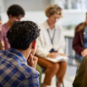A group of people sit in a circle of chairs as one man props his chin on his hand