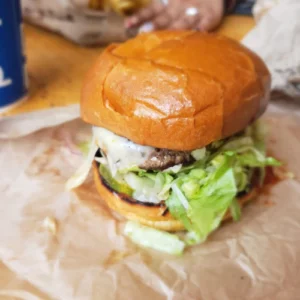 A cheese burger with lettuce sits on paper with a cup in the background