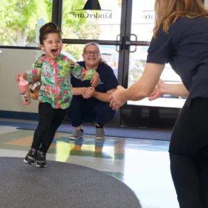 A happy child runs from a crouching woman into the arms of a standing woman viewed from behind